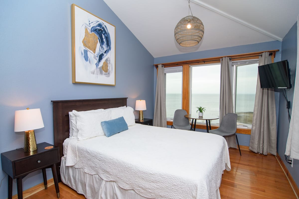 A hotel room with a large bed, two bedside tables with lamps, a TV, a small table with a chair, and a large window with curtains.