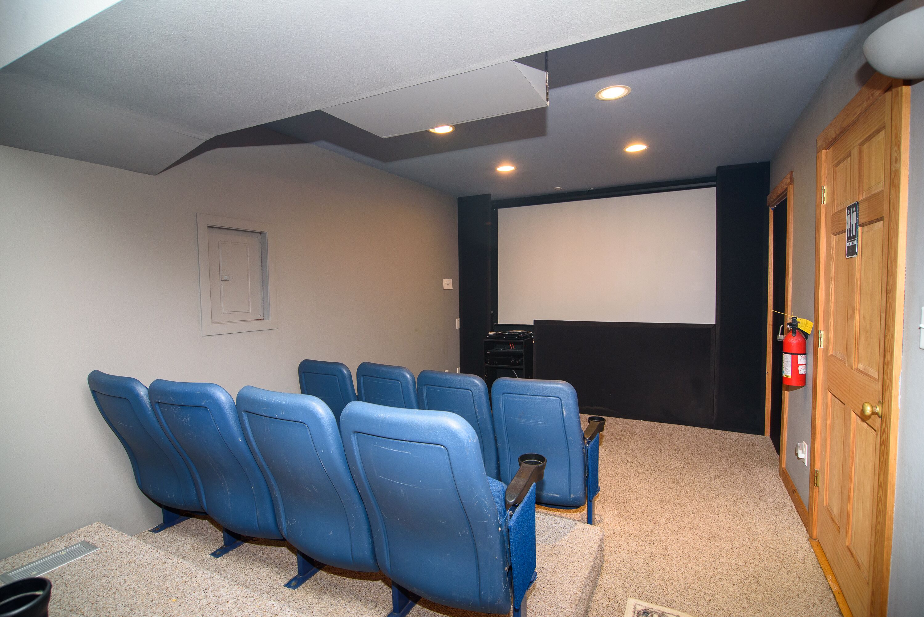 A home theater room with six blue chairs, a carpeted floor, a white screen, and a wooden door.