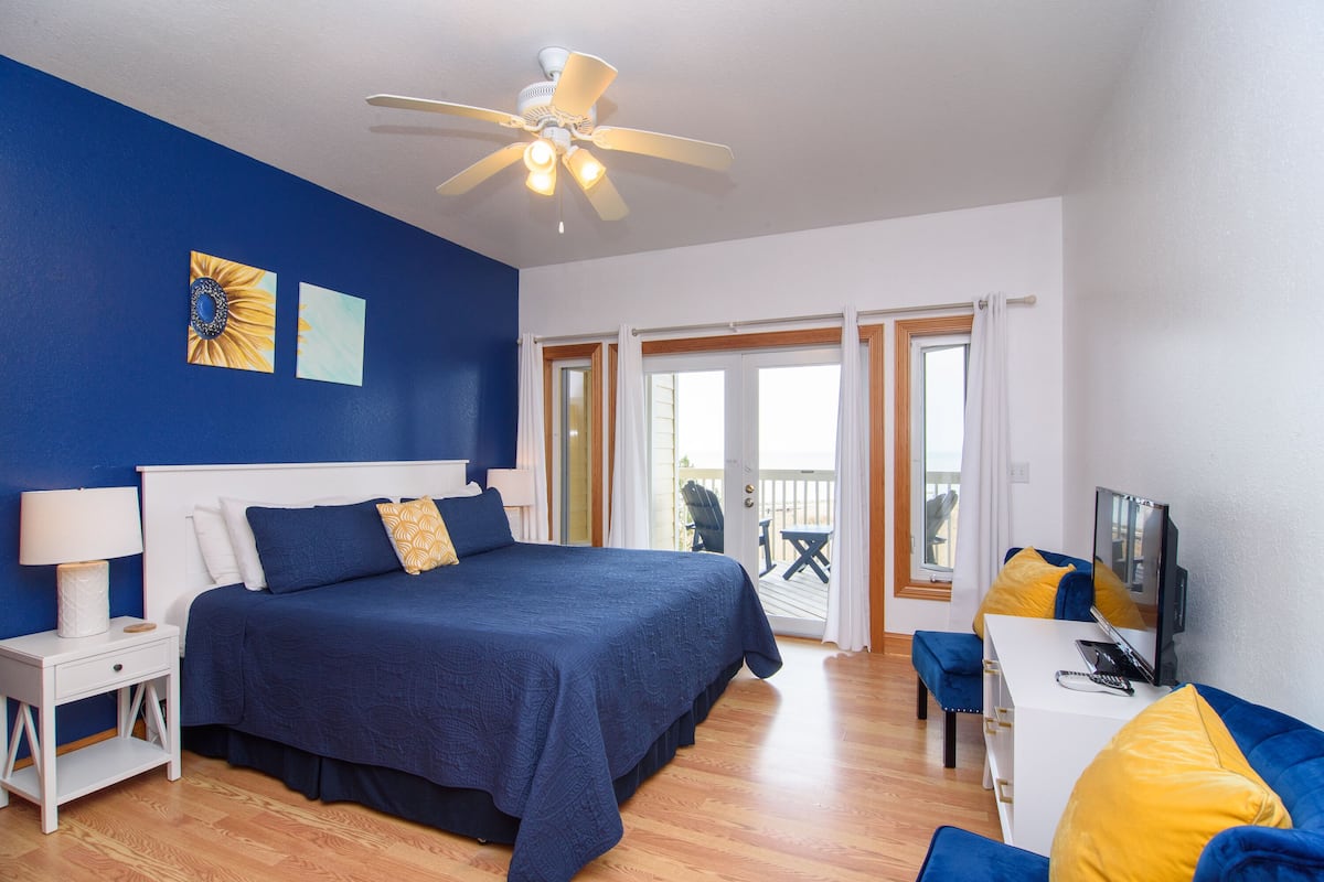 A bedroom with a large bed, a ceiling fan, a desk with a TV, and a balcony with a chair.