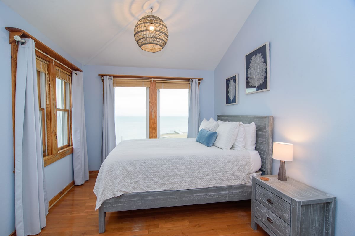 A bedroom with a bed, wooden nightstand, and a view of the sea.