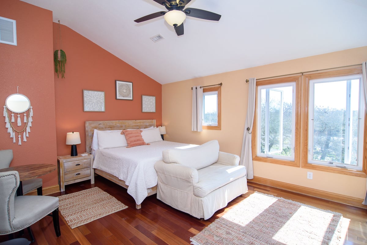 A bedroom with a bed, a chair, a desk, a nightstand, a ceiling fan, and large windows.