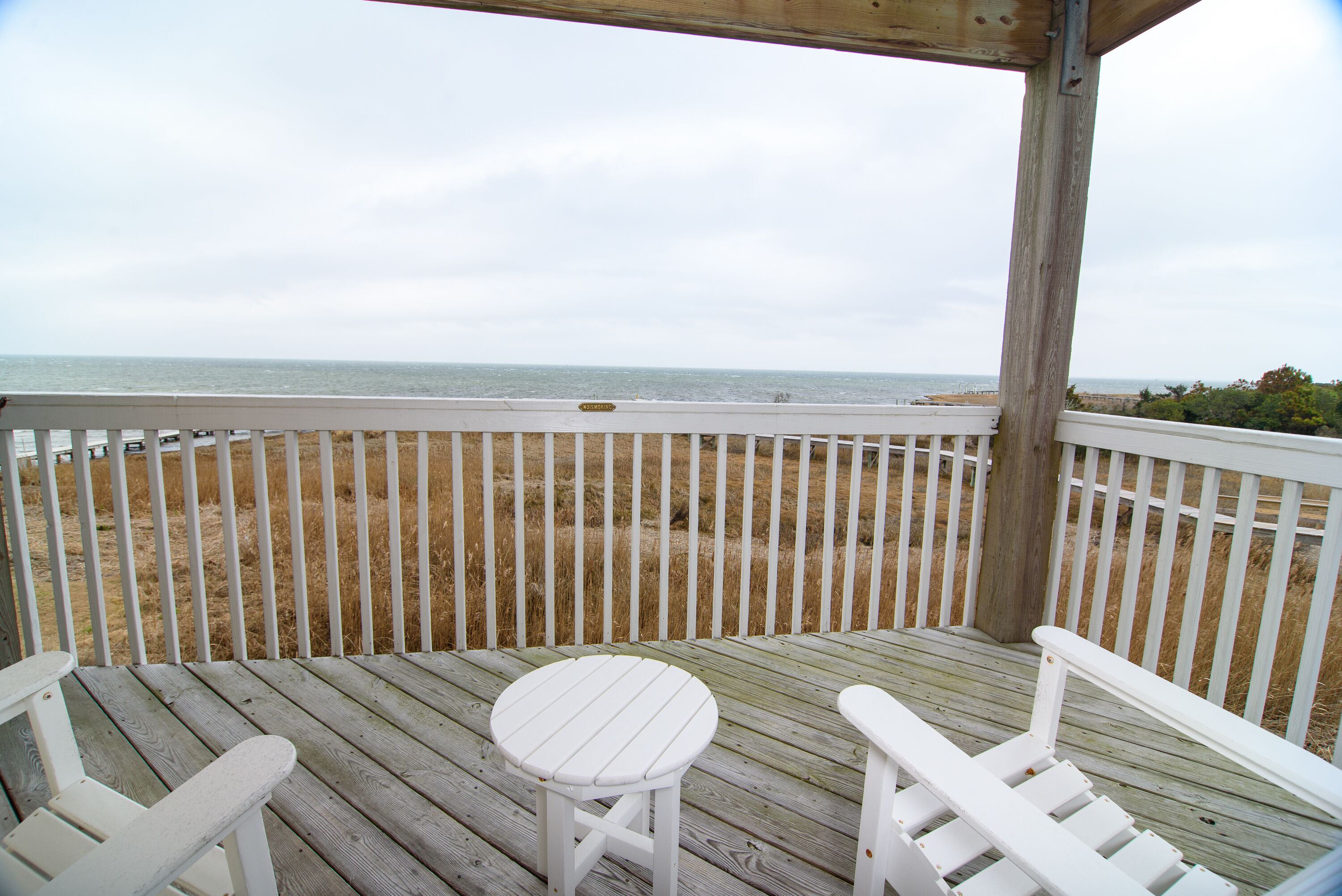 A wooden deck with white railings and two white chairs facing a beach.