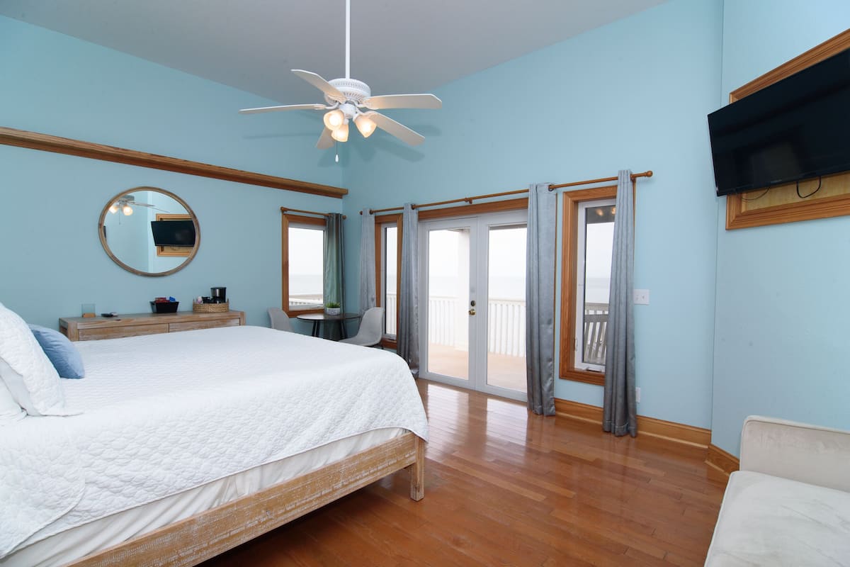 A bedroom with a bed, a ceiling fan, a TV, a window with curtains, and a balcony.