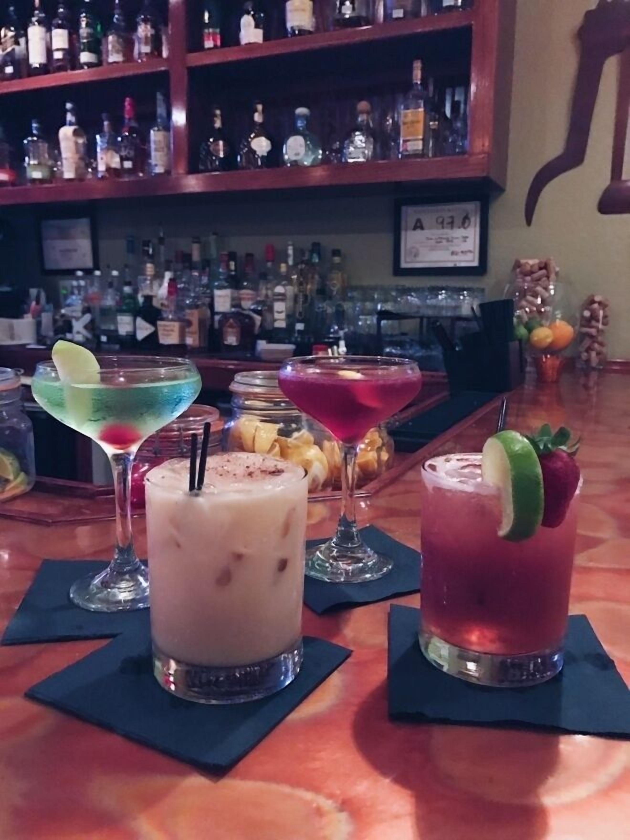 A bar area with various cocktails on the counter and a shelf of liquor bottles in the background.