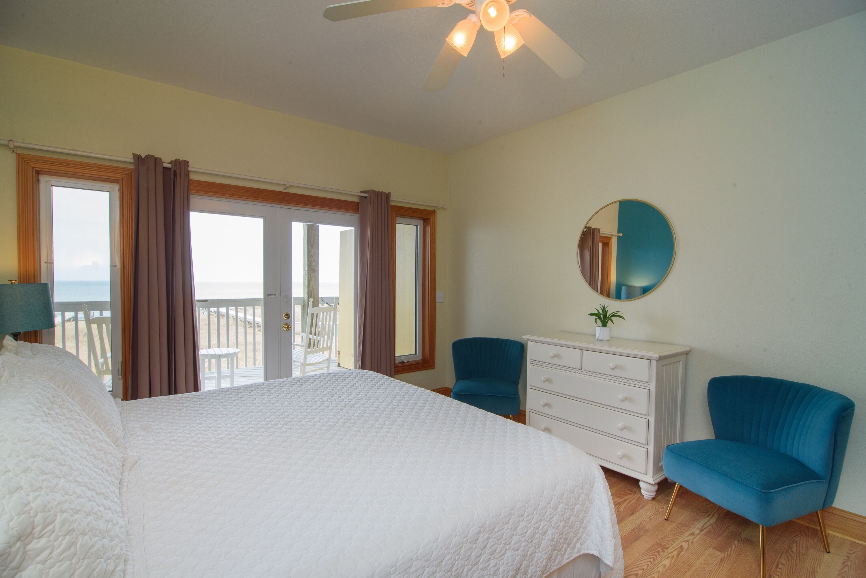 A bedroom with a large bed, a ceiling fan, a dresser, two chairs, a round mirror, and a view of the beach through the window.