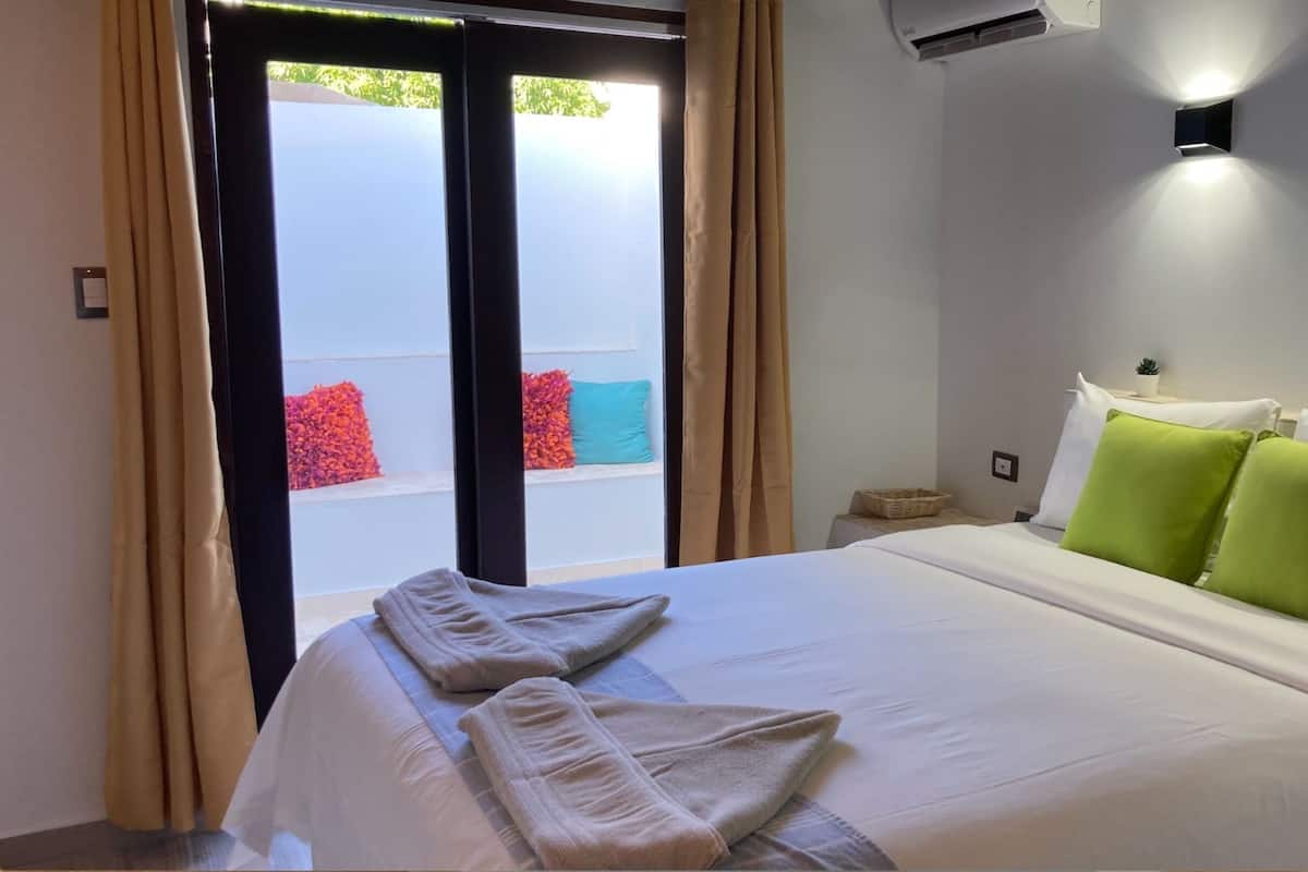 Habitación de hotel con una cama, dos toallas, una mesita con una planta y una ventana con cortinas.