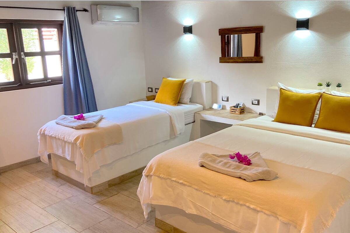 Habitación de hotel con dos camas, cada una con ropa blanca y almohadas amarillas, una mesita de noche pequeña, aire acondicionado instalado en la pared y una ventana con cortinas.