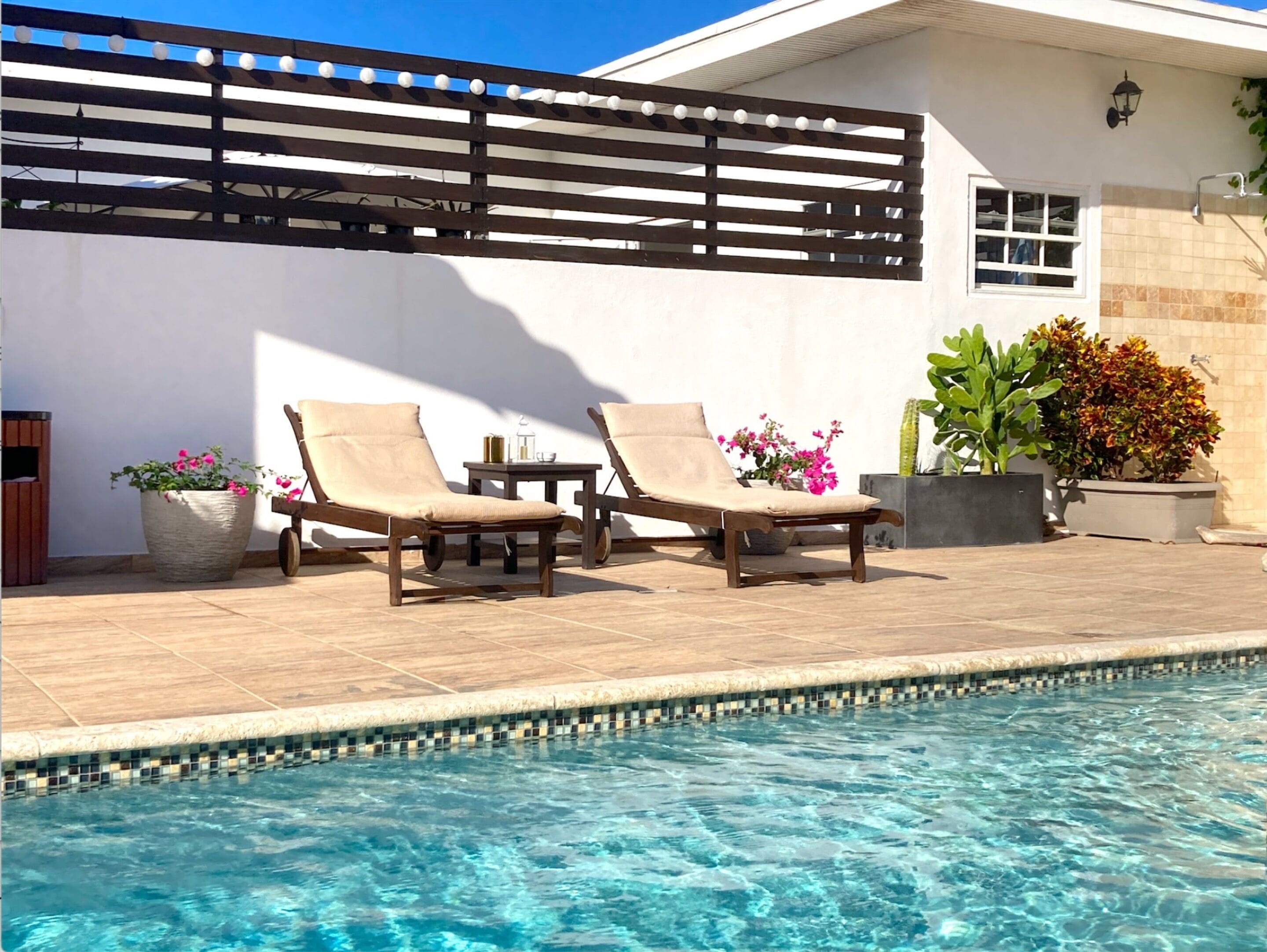 Ένας χώρος δίπλα στην πισίνα με δύο ξαπλώστρες, ένα μικρό τραπέζι και γλάστρες με φυτά.
