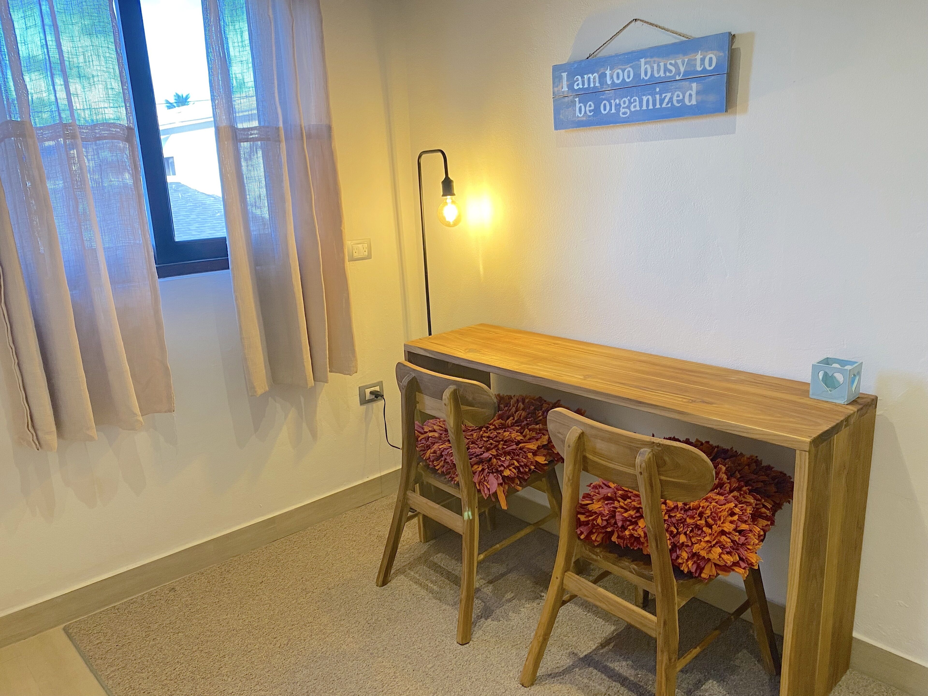 Une petite pièce avec un bureau en bois et deux chaises, un luminaire fixé au mur, et une pancarte indiquant : « Je suis trop occupé(e) pour être organisé(e). »