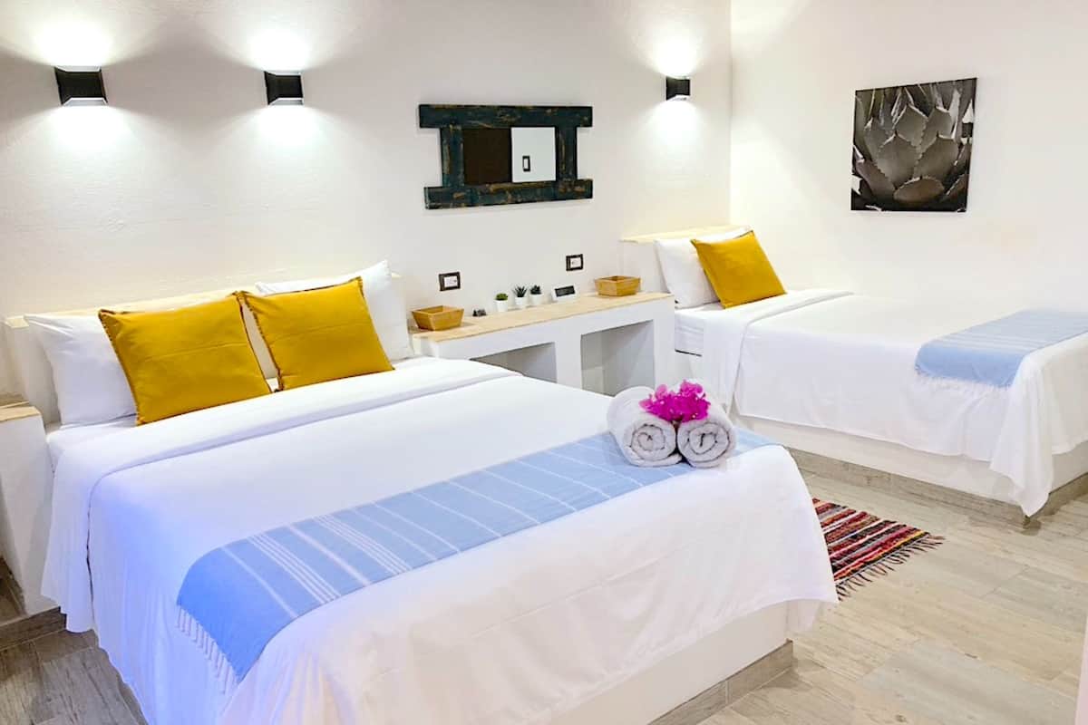 Habitación de hotel con dos camas, cada una con ropa blanca y almohadas amarillas. Hay un escritorio pequeño con una lámpara, un espejo y algunos objetos decorativos. Una alfombra colorida cubre el suelo.