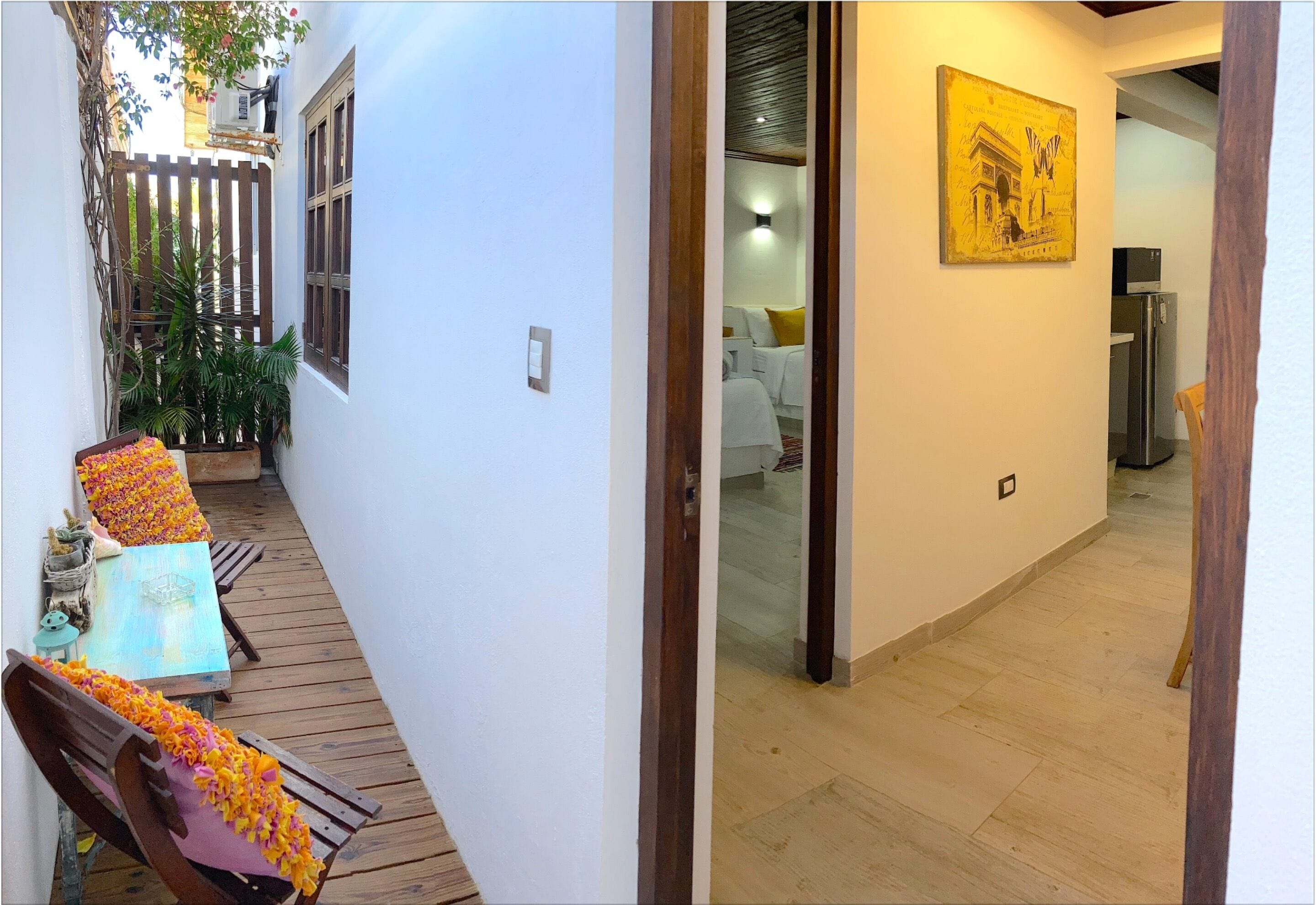 A narrow corridor with a wooden bench and a potted plant on the left, a bed with white bedding and a yellow pillow in the center, and a refrigerator in the background on the right.