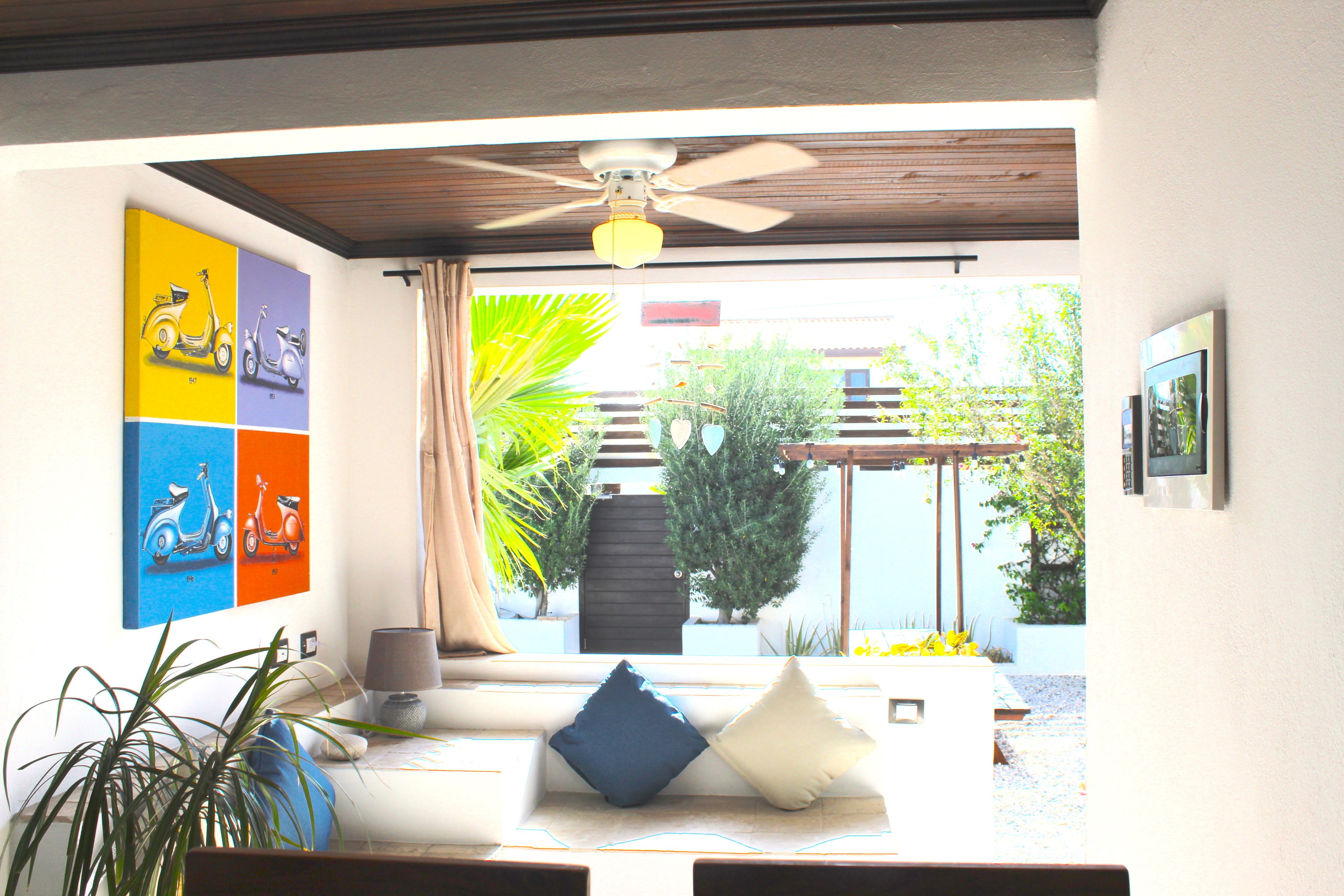 Un salon moderne doté d’un ventilateur de plafond, d’un canapé avec des coussins, d’une table basse et offrant une vue sur le jardin extérieur.