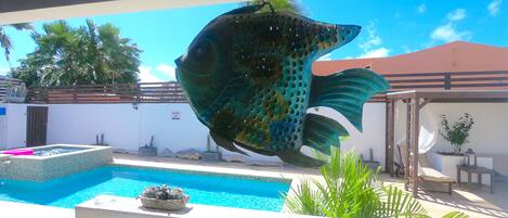 A swimming pool with a large fish sculpture, a covered patio area, and a view of a building with a red roof.