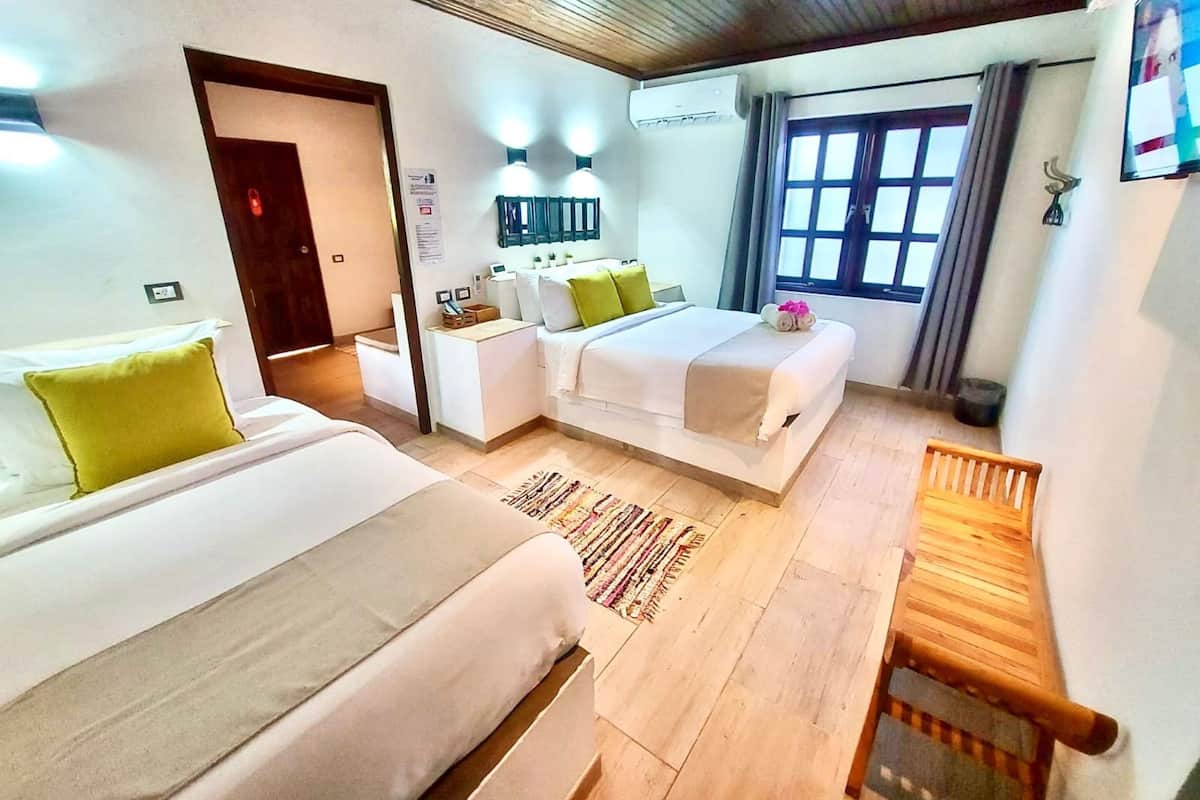 Habitación de hotel con dos camas, un sofá, un banco de madera, una ventana con cortinas y un equipo de aire acondicionado.