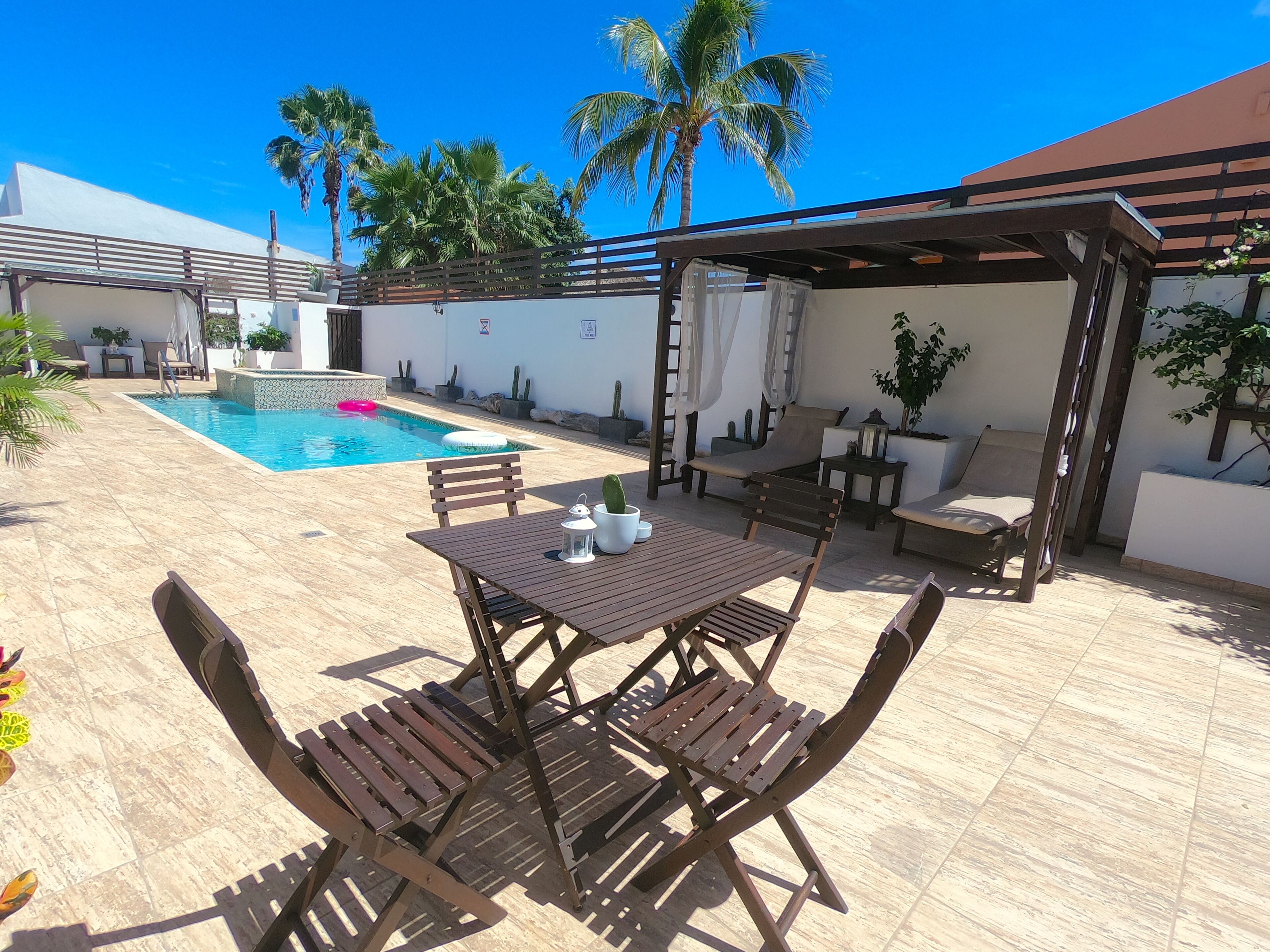 Zona de piscina con mesa y sillas, rodeada de palmeras y una pérgola.