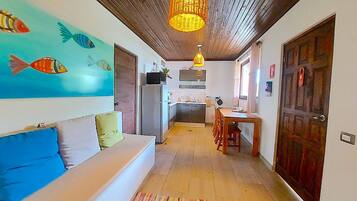A cozy interior with a wooden ceiling, a colorful fish mural, a white sofa with cushions, a kitchen area with a refrigerator, and a dining table with chairs.