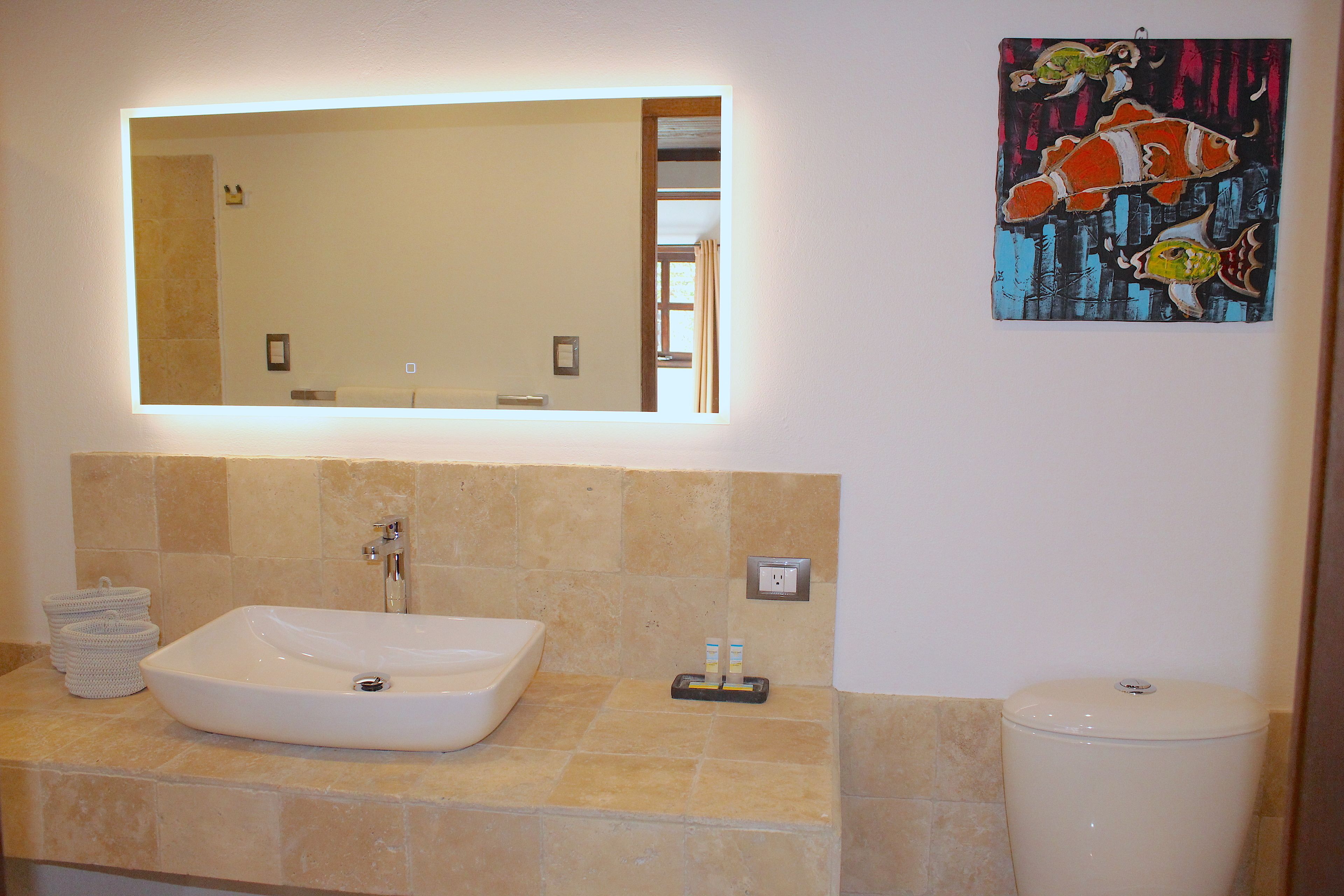 Een badkamer met een grote spiegel, een wit wastafelmeubel en een toilet. Op de muur hangt een schilderij en er is een klein plankje met toiletartikelen.