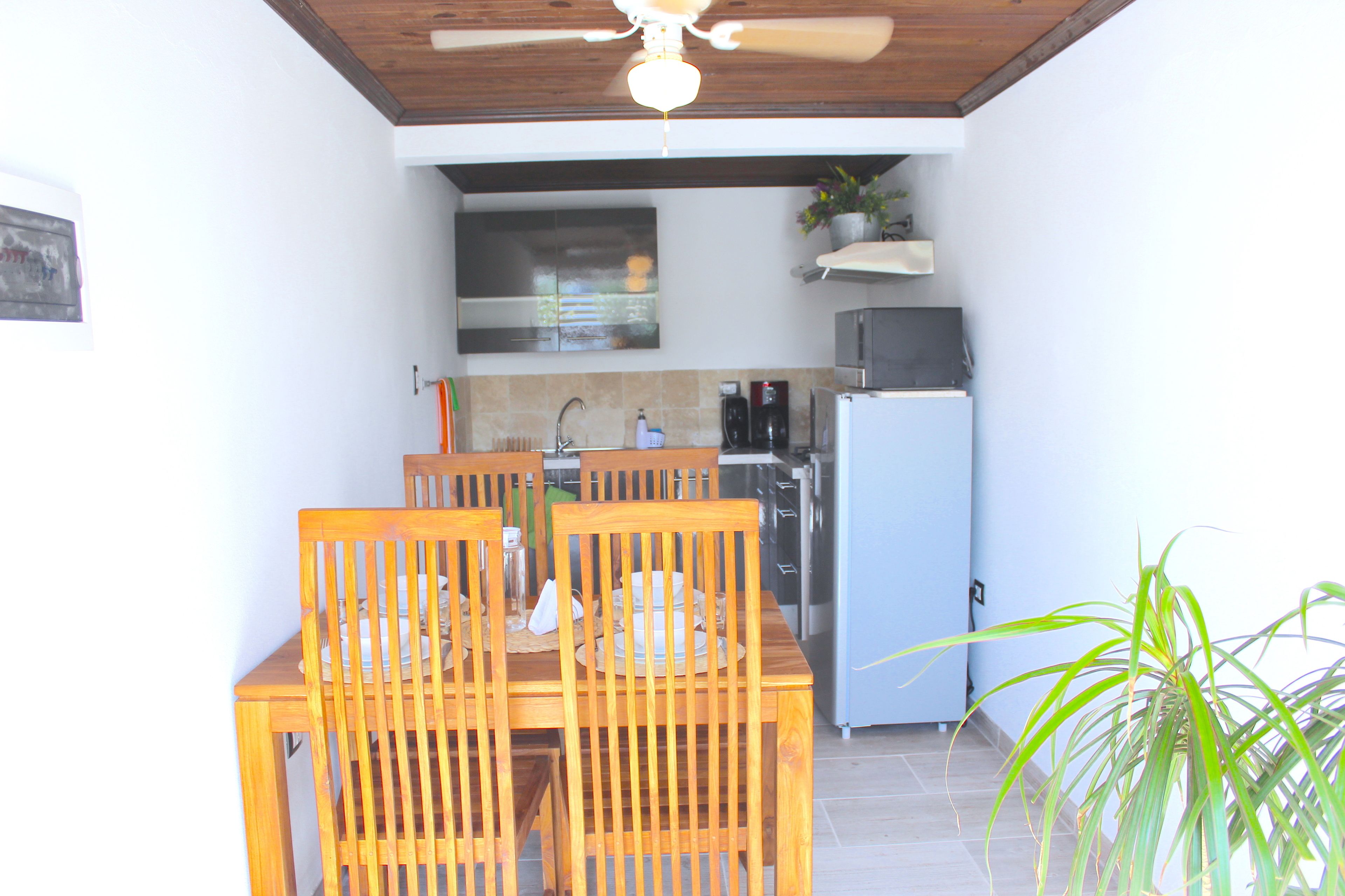 Un comedor con una mesa y sillas de madera, un ventilador de techo y una cocina al fondo.