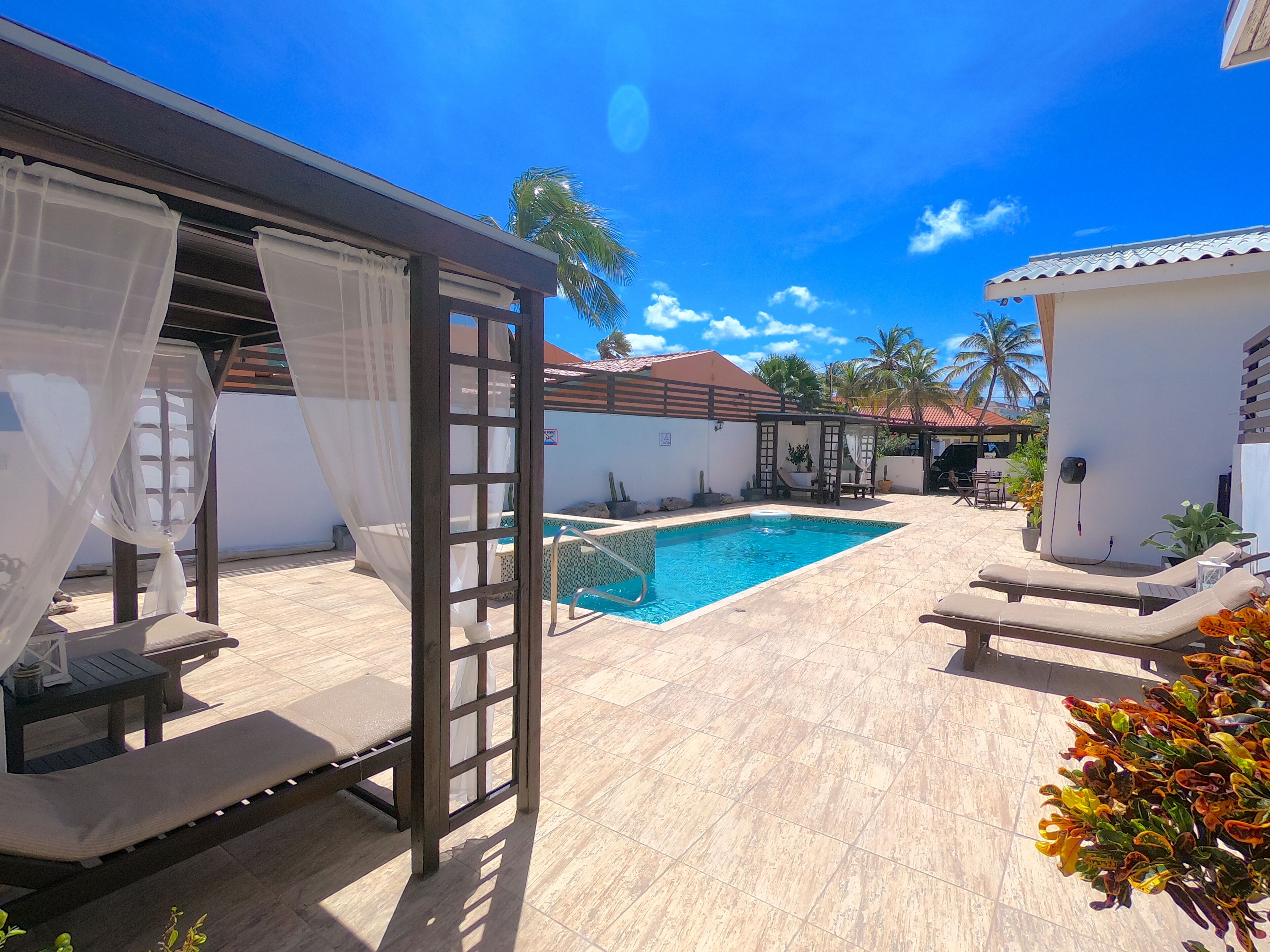 Una piscina con un pérgola y sillones de descanso, rodeada por un patio de baldosas y palmeras.