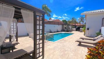 Una piscina con un pérgola y sillones de descanso, rodeada por un patio de baldosas y palmeras.