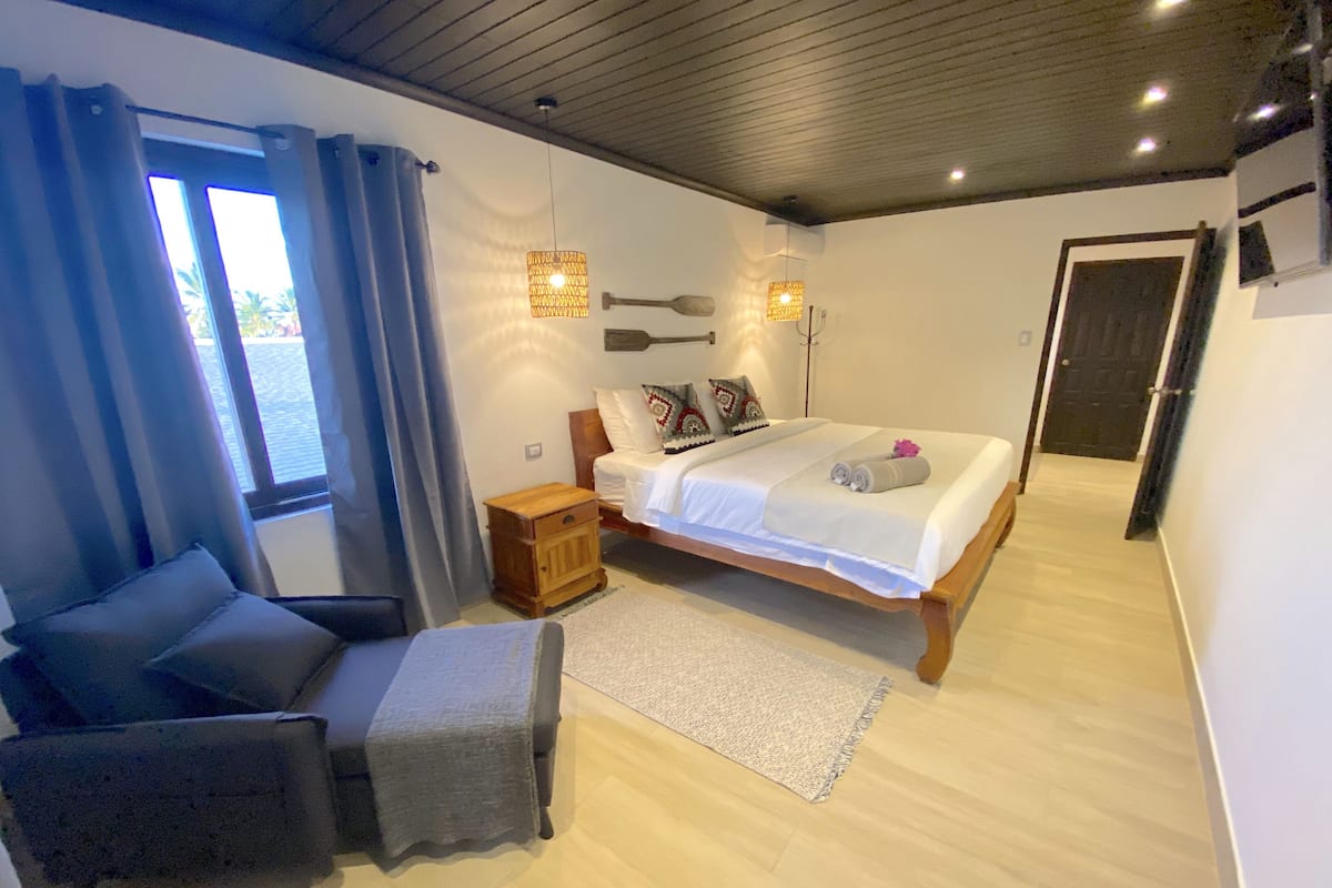 Un dormitorio con cama, mesitas de noche, una silla, una ventana con cortinas y una puerta.