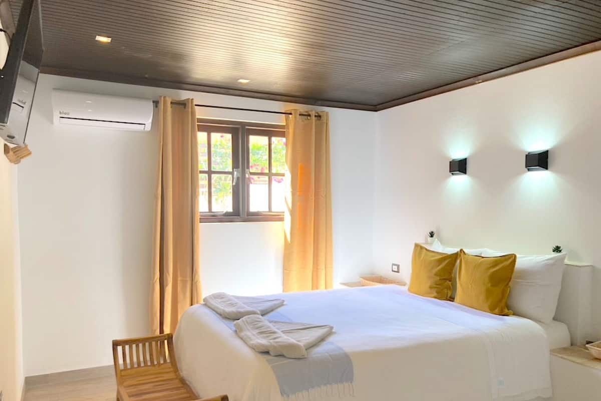 Una habitación de hotel con una cama, un banco de madera, una ventana con cortinas y un aire acondicionado de pared.