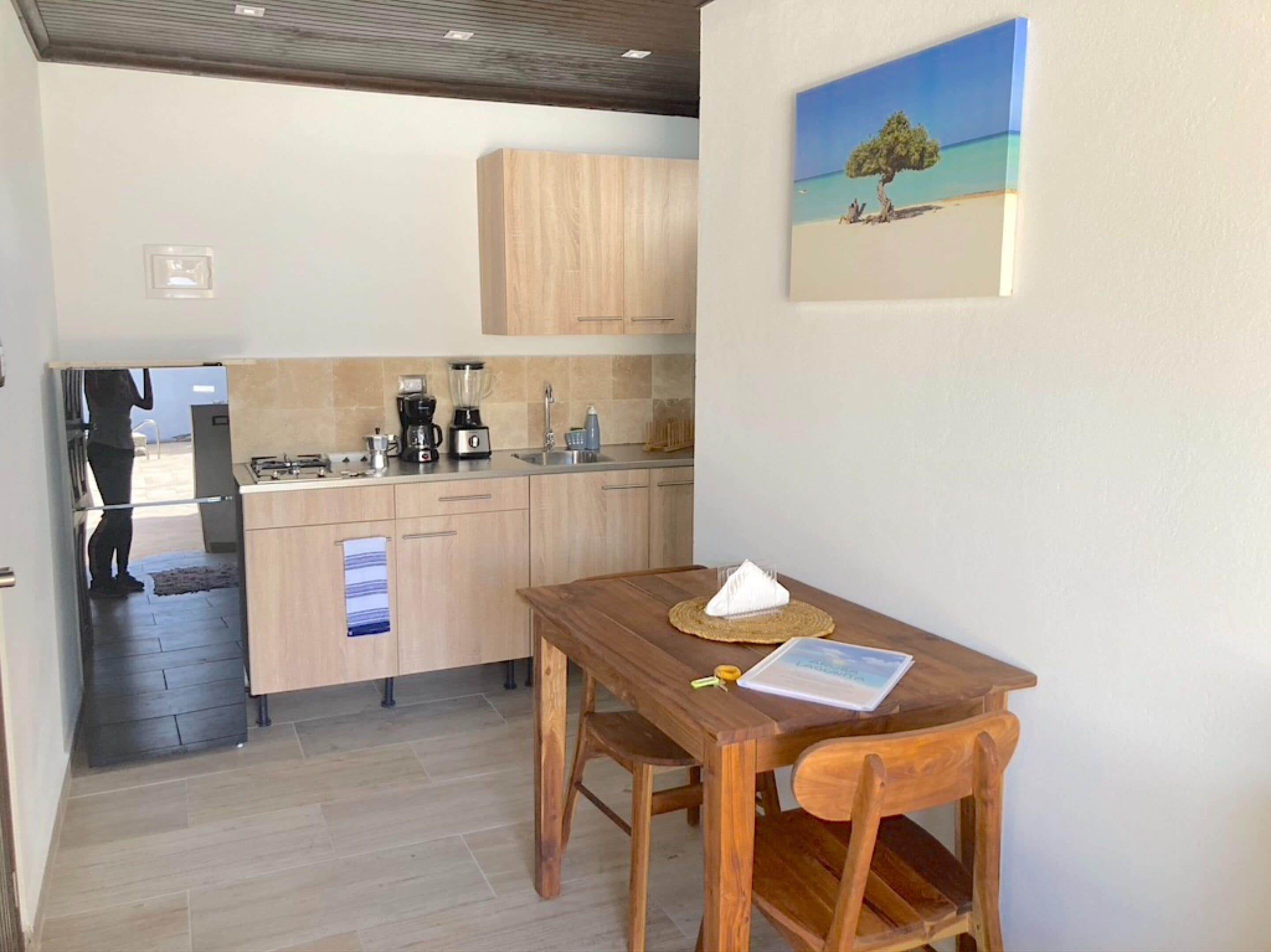 A compact kitchen with wooden cabinets and a dining table with chairs. A painting of a tree by the sea hangs on the wall.