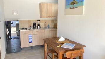 A compact kitchen with wooden cabinets and a dining table with chairs. A painting of a tree by the sea hangs on the wall.