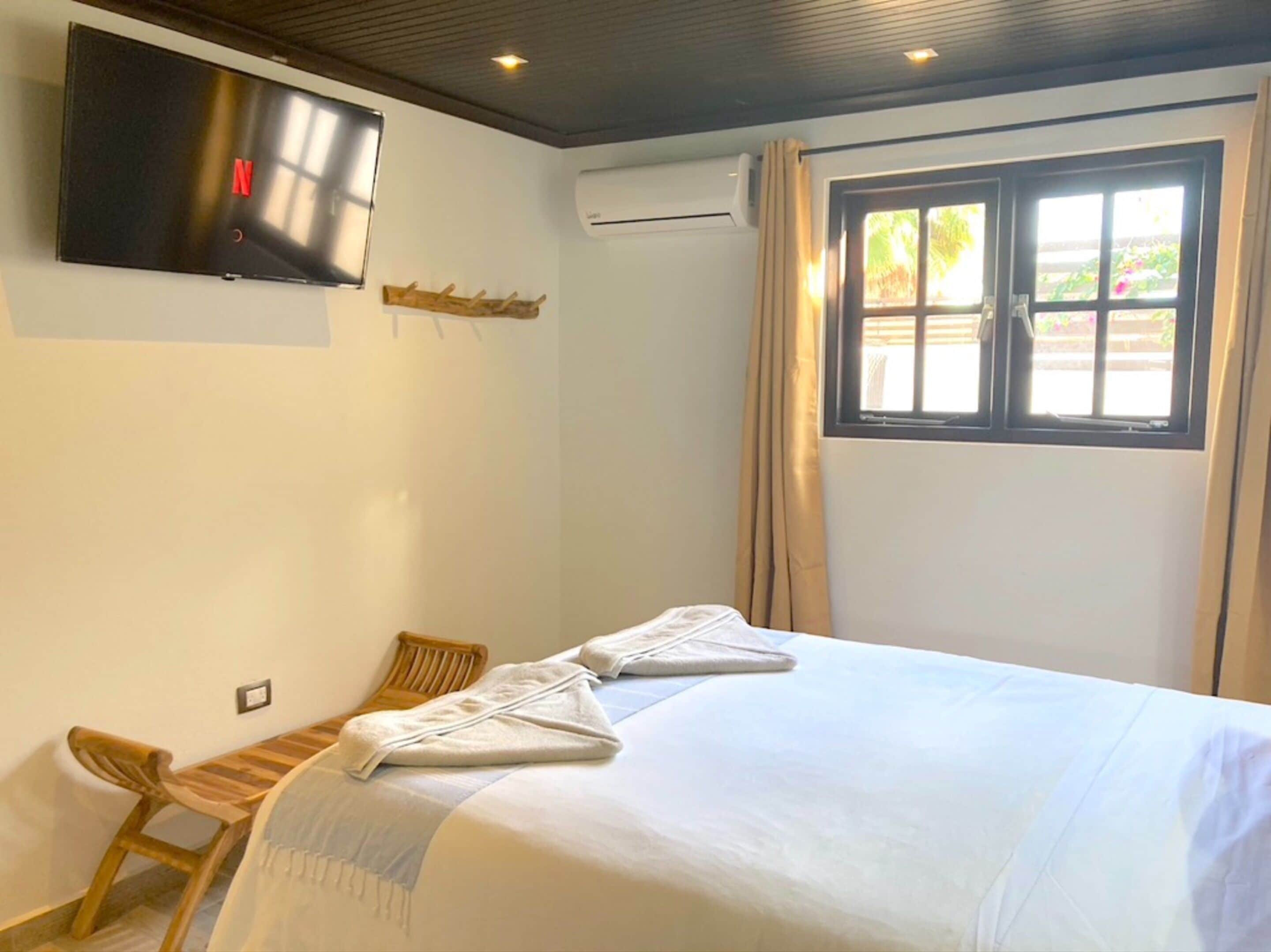 Habitación de hotel con cama, televisor montado en la pared, ventana con cortinas y una silla de madera.