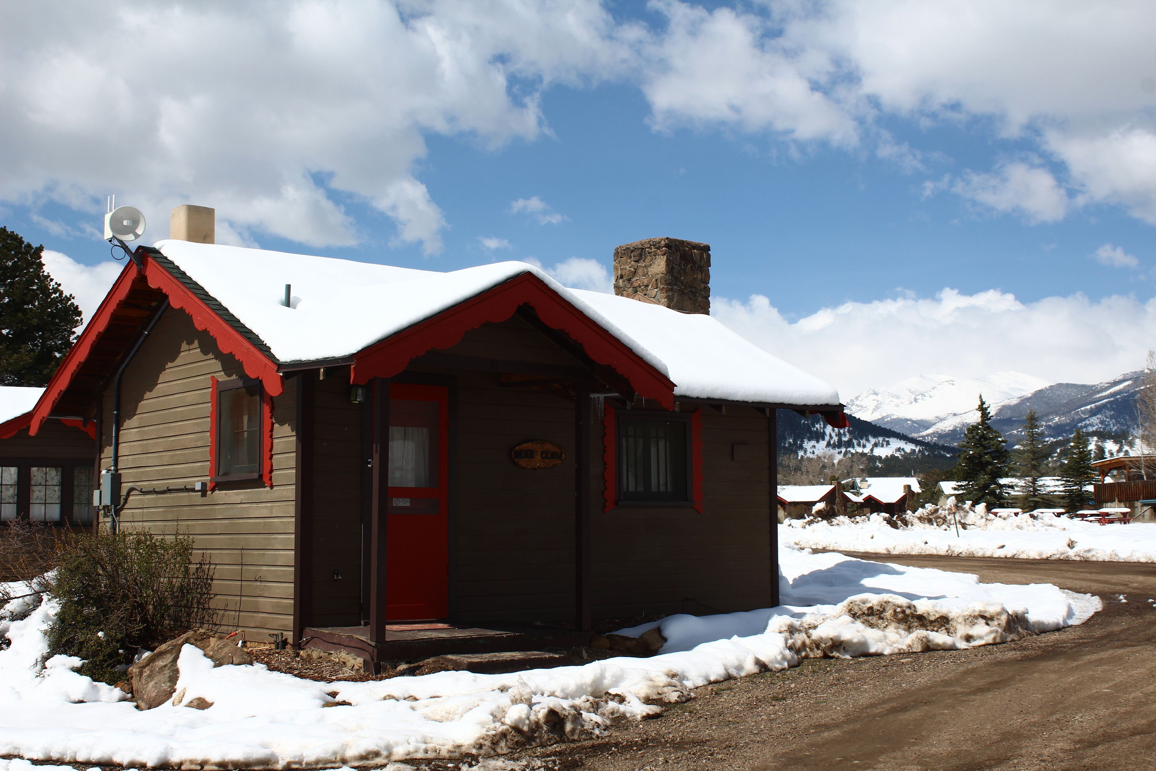 A small cabin with a red door and a chimney, surrounded by snow and mountains in the background.