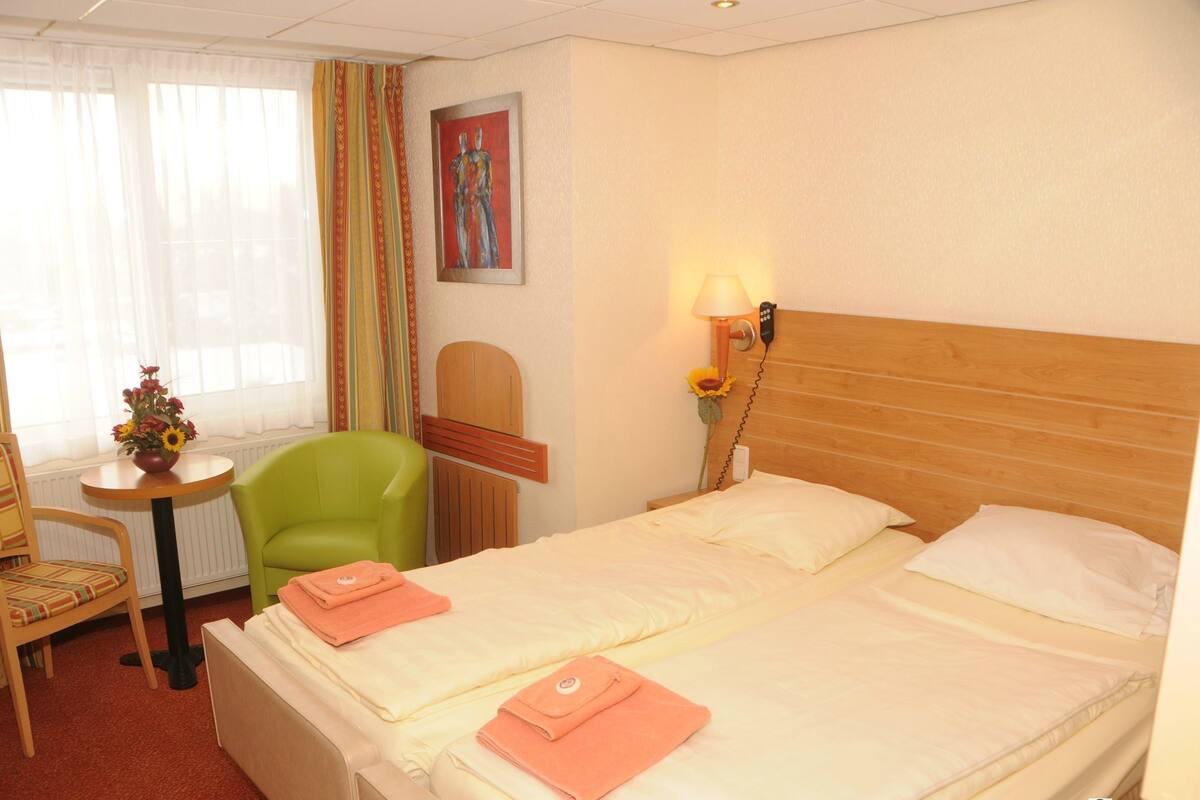 A hotel room with two beds, a chair, a small table with a vase of flowers, and a painting on the wall.