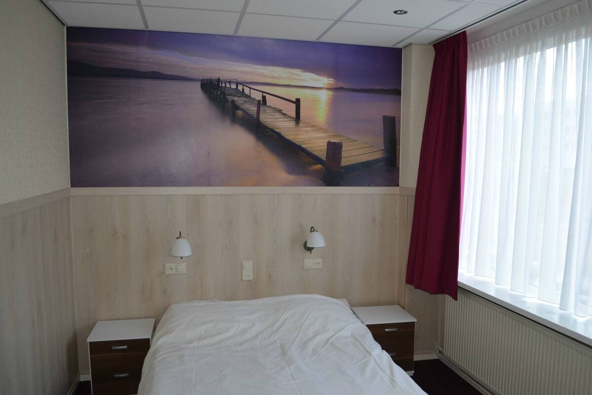 A hotel room with a bed, a nightstand, a wall-mounted lamp, a window with curtains, and a mural of a sunset over a pier.