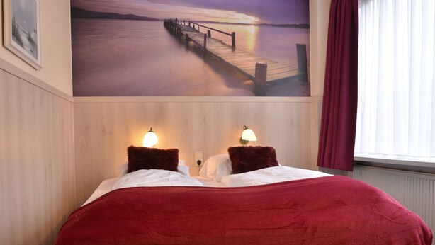 A hotel room with a bed, two wall-mounted lamps, a large painting of a pier, and a window with curtains. - Hotel Het Galjoen (Dronten)