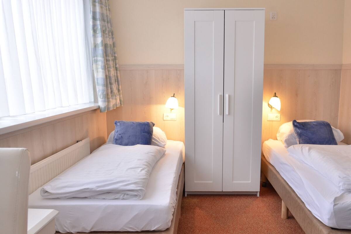 A room with two single beds, each with a pillow and a white duvet. There is a wardrobe with two doors and a light fixture above it.