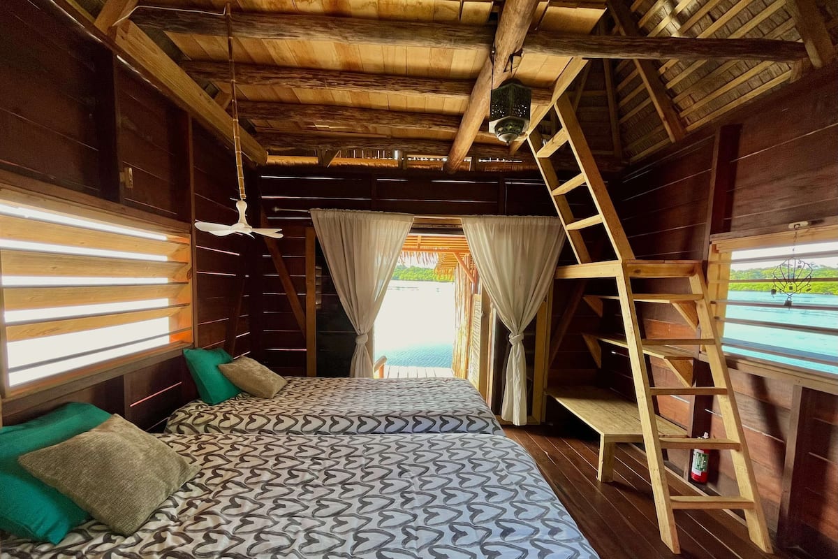 Une pièce de cabane en bois comprenant un lit, des oreillers, une échelle et donnant sur un plan d’eau.
