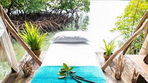 Een houten terras met een bed, omringd door natuurlijke elementen zoals bomen en planten, met uitzicht op een rustig waterlichaam.