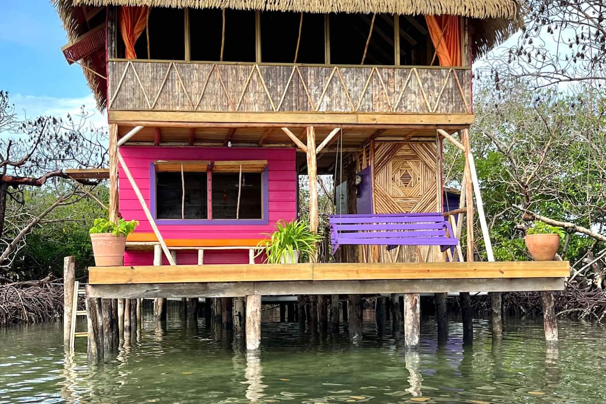 Une maison à deux étages, construite sur pilotis, avec un toit de chaume, des poutres en bois et un balcon agrémenté de plantes en pot.