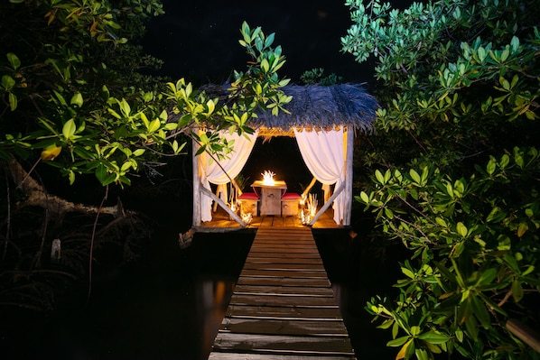 A wooden pathway leading to a thatched-roof structure with white curtains and a small table with a light source.