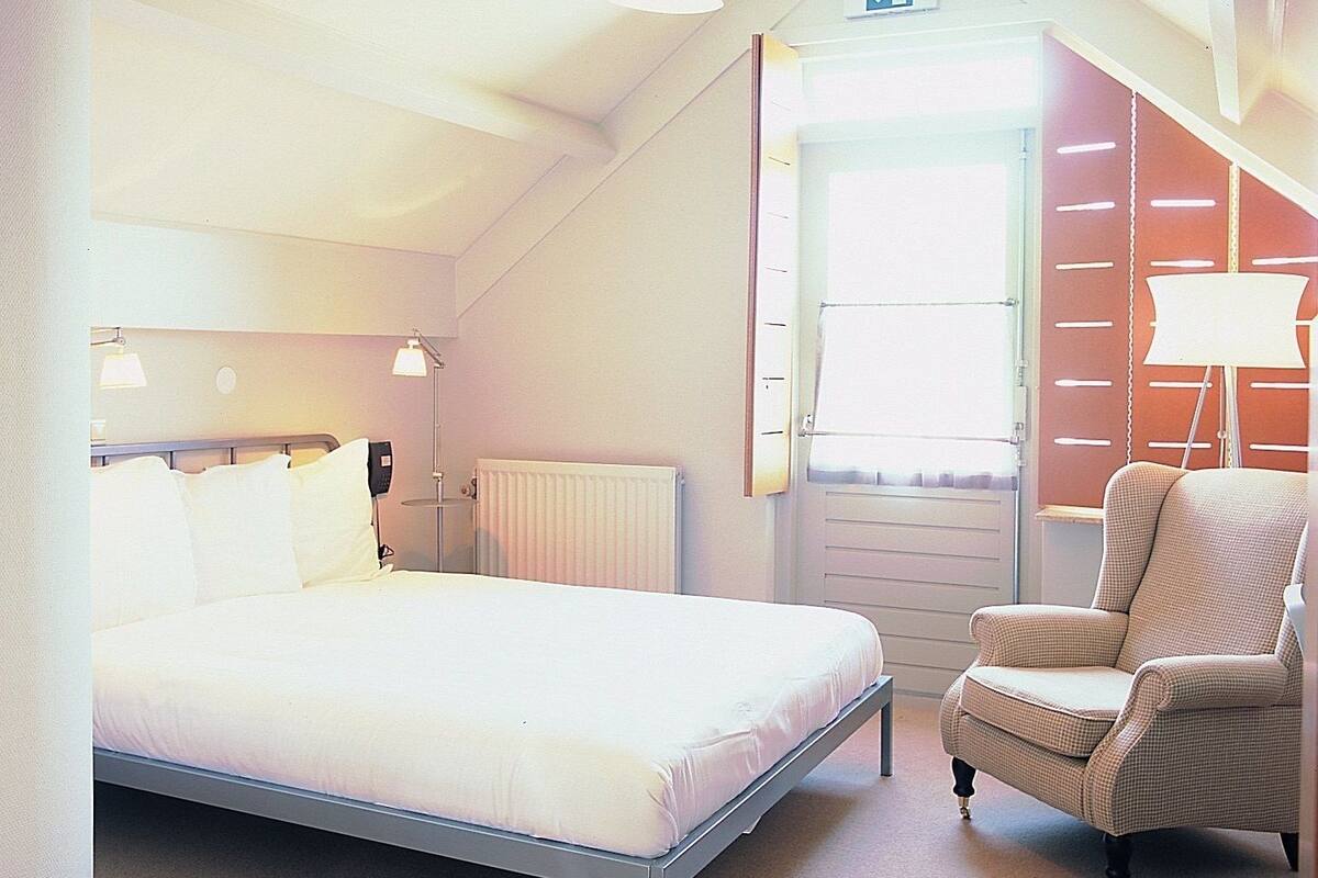 Habitación de hotel con cama individual, lámpara de noche, silla y ventana con persianas.