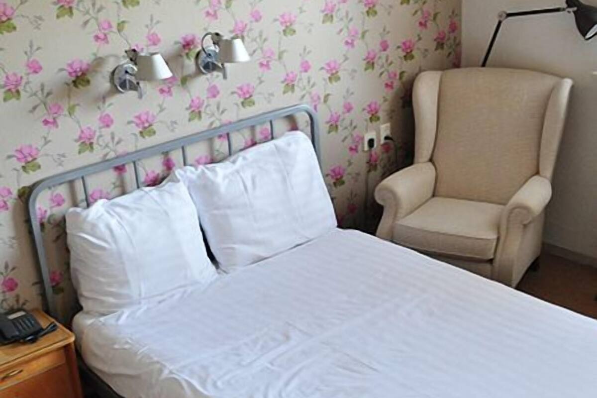 Una cama bien hecha con sábanas blancas, cabecera de metal y un estampado de papel tapiz floral.