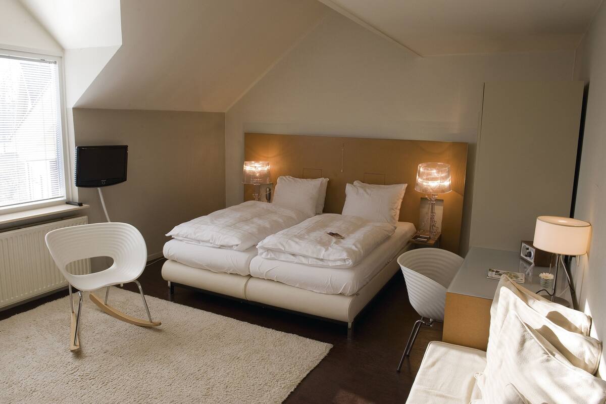 Un dormitorio con cama, lámparas de noche, una silla y un escritorio con lámpara.