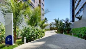 Um caminho ladeado por vegetação exuberante e edifícios modernos, com uma placa indicando 'Praia da Jatiba' e 'Saida Avenida Álvarez Otacílio'.