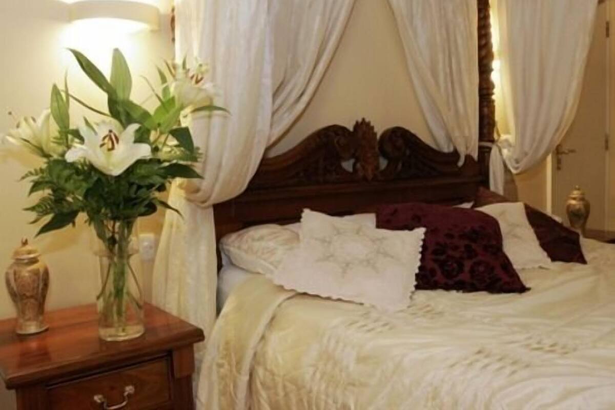 A bedroom with a four-poster bed, white bedding, a vase of flowers on a nightstand, and a lamp.