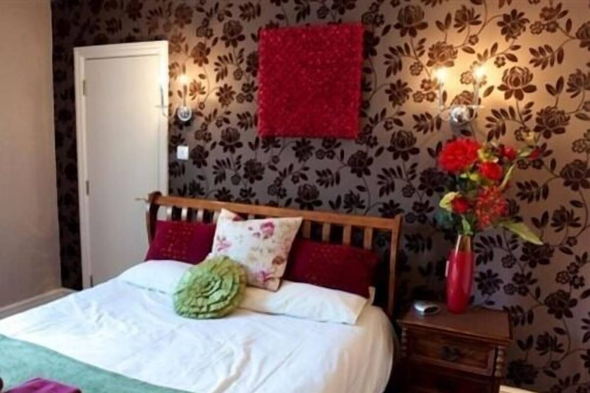 A bedroom with a wooden bed, white bedding, a green pillow, red and floral pillows, a red vase with flowers, and a wall with floral wallpaper.