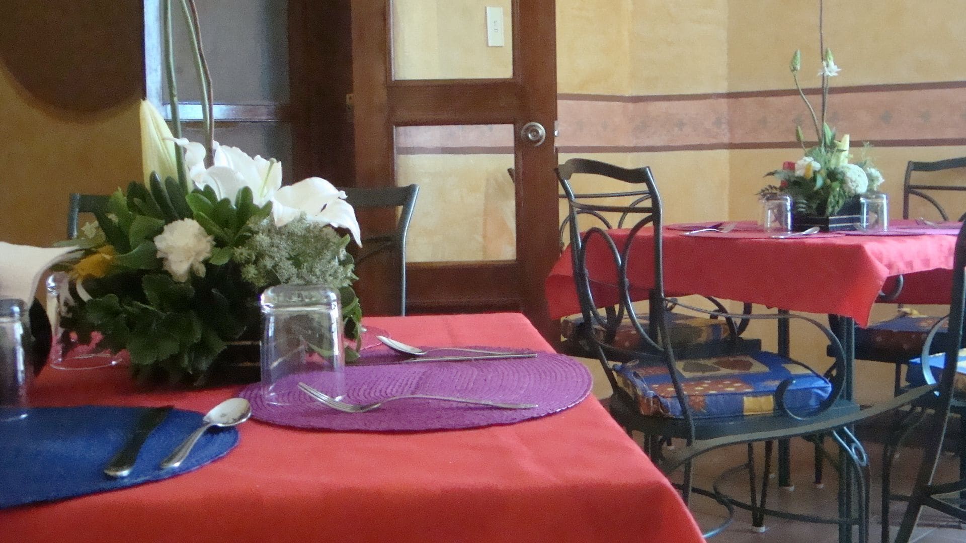 Un comedor con mesas dispuestas para dos, con manteles rojos, posavasos morados y centros de mesa florales.