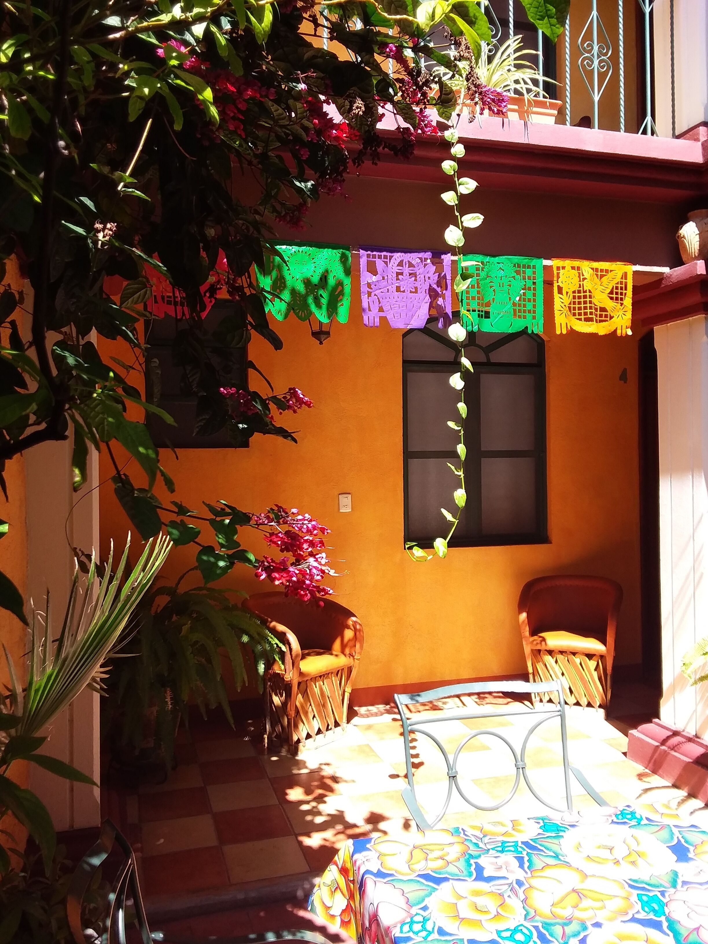 Una habitación con piso a cuadros, una cama con colcha floral de colores, dos sillas de mimbre y una ventana con marco negro.