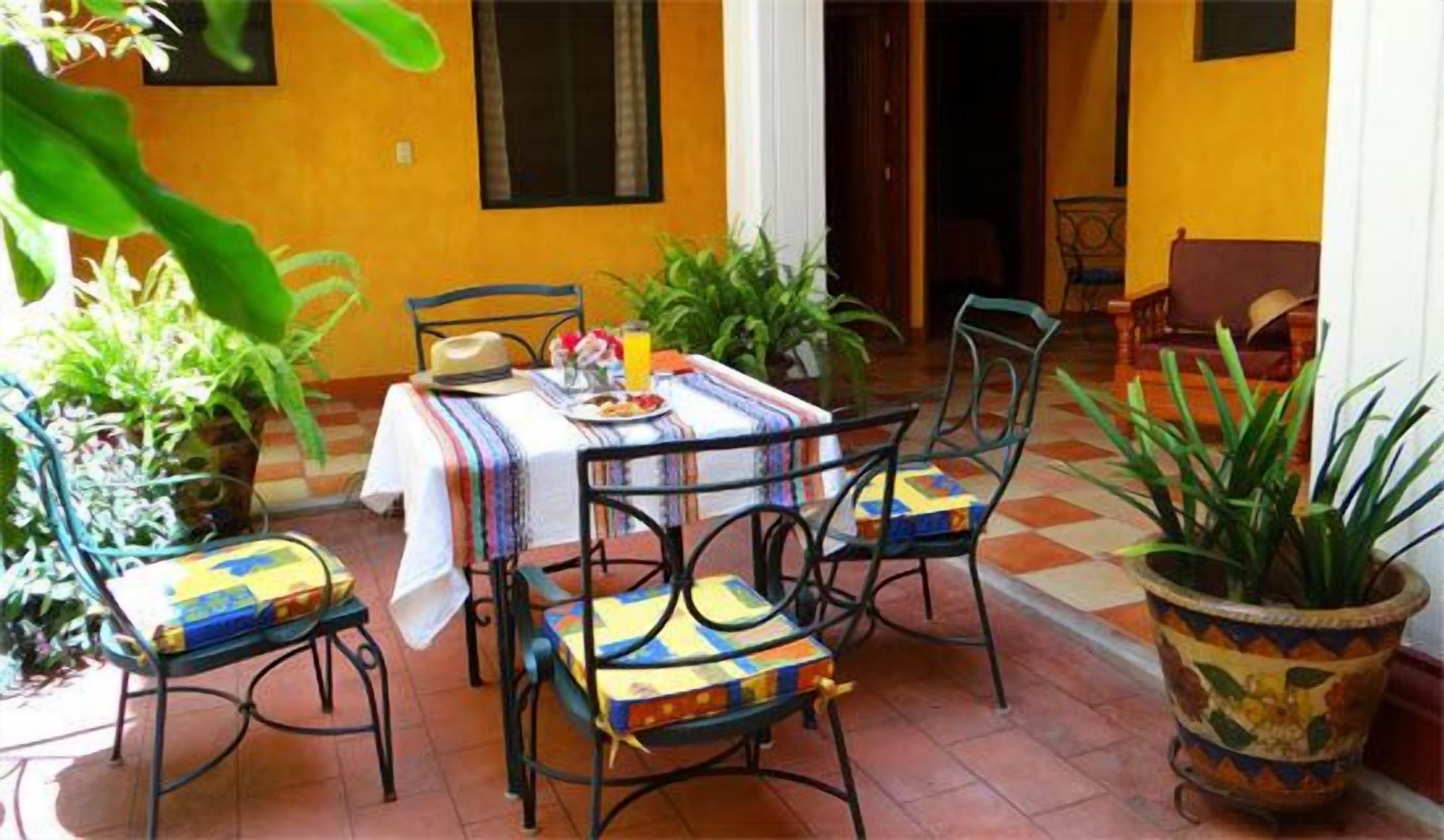 An outdoor patio with a table set for two, surrounded by chairs with colorful cushions. There are potted plants and a tiled floor.