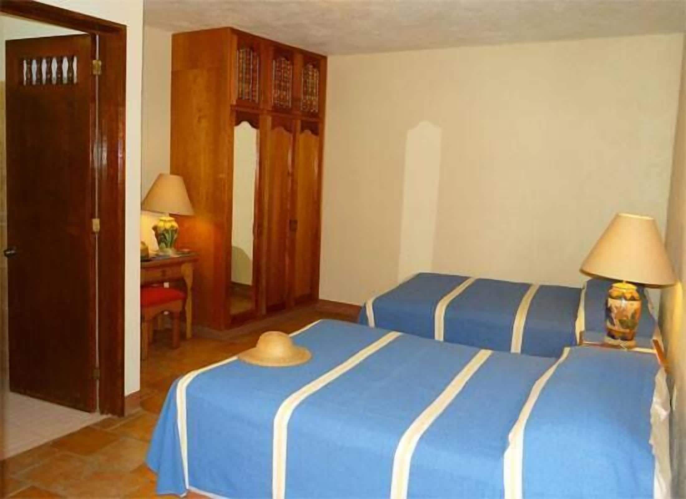 A hotel room with two beds, wooden wardrobe, and a small table with a lamp.