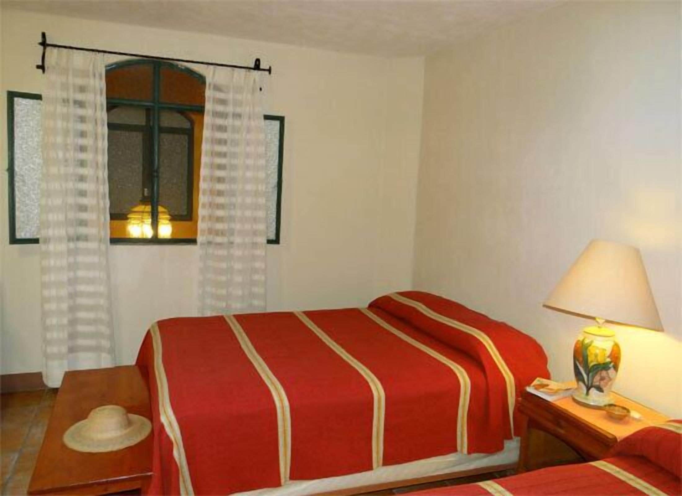 Una habitación de hotel con una cama, una mesita de noche con lámpara, una ventana con cortinas y un sombrero sobre una silla.