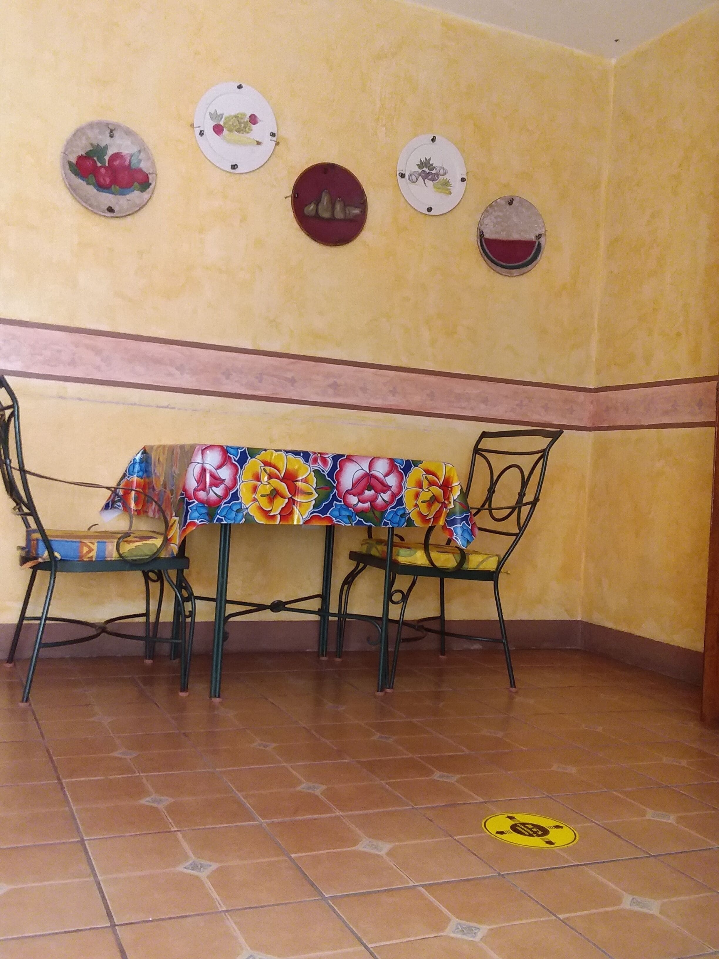 A dining area with a table covered by a colorful tablecloth, two chairs, and a wall decorated with plates.